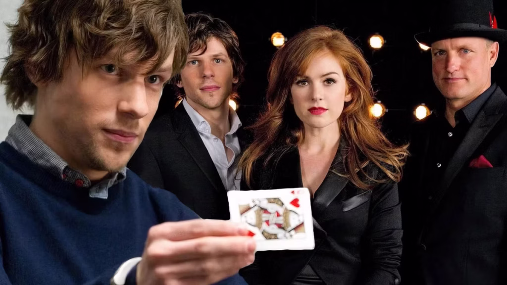 A promotional image from Now You See Me showing four magicians standing together. In the foreground on the left, a young man with tousled hair holds up a playing card (the Queen of Hearts) toward the camera. Behind him stand three other characters: a man with short dark hair in a suit, a woman with long red hair wearing a black jacket, and a man in a dark suit and black hat with a red feather. Warm stage lights glow behind them, giving the scene a dramatic, theatrical feel.
