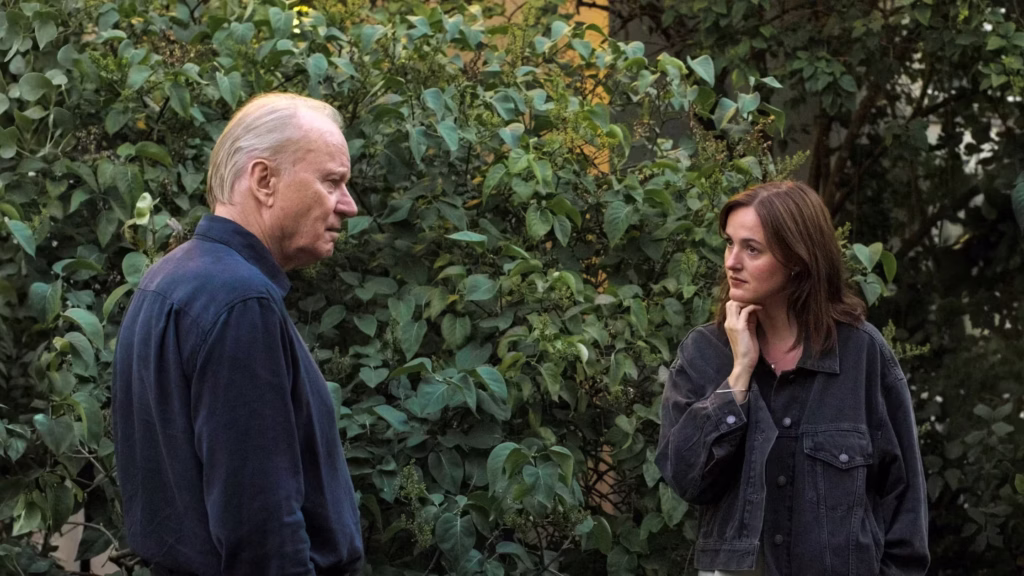 An older man with thinning blond hair and a blue shirt stands in profile, looking toward a younger woman with shoulder-length brown hair. The woman, wearing a dark denim jacket, rests her chin on her hand and looks back at him with a serious expression. They are positioned outdoors in front of a dense wall of green leafy bushes.