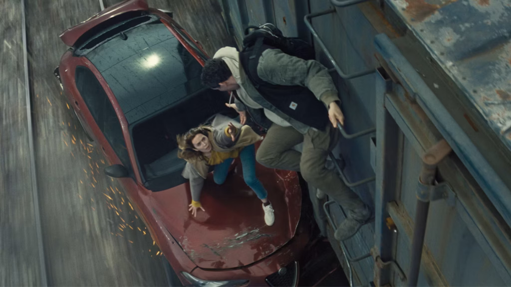 An overhead medium shot captures a high-stakes action sequence on a moving train. On the left, a damaged red Alfa Romeo sedan scrapes alongside a rusty blue cargo train car, sending sparks flying onto the grey tracks below. A woman with light brown hair, wearing a yellow top, blue jeans, and a grey jacket, clings to the bonnet of the car while stretching her left hand upward. A man in a grey hoodie, black jacket, and khaki trousers hangs precariously from the metal ladder on the side of the blue train car, leaning down to grasp her outstretched hand.